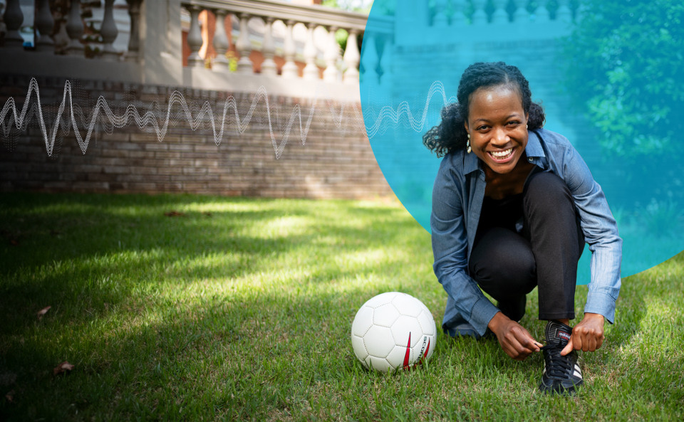 A young woman kneels while tying her shoe. A soccer ball sits on the grass beside her. An overlay signifying a brainwave is behind her.